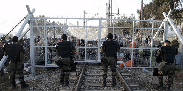 Macedonian policemen stand in front of a gate over rail tracks as migrants wait behind at the Greek-Macedonian border
